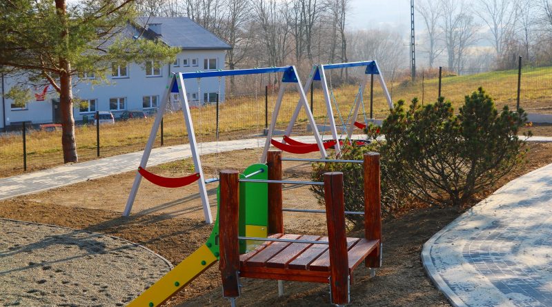 Na zdjęciu widoczne dwie hustawki oraz zjeżdzalnia, które stanowia wyposażenie placu zabaw dla dzieci.