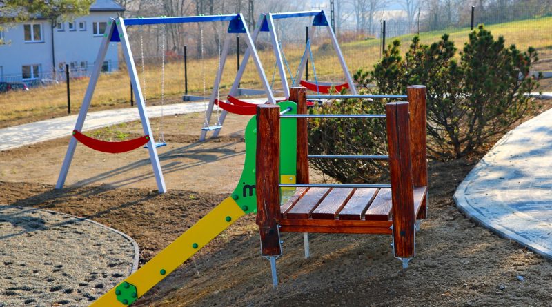 Na zdjęciu widoczne dwie hustawki oraz zjeżdzalnia, które stanowia wyposażenie placu zabaw dla dzieci.